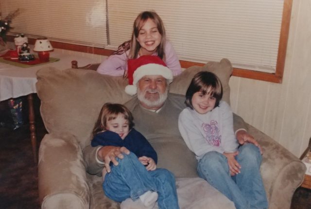 A senior man wearing a Santa hat sits with a grandchild in each arm and one standing behind the chair. One strikes a silly pose, all smile joyfully.